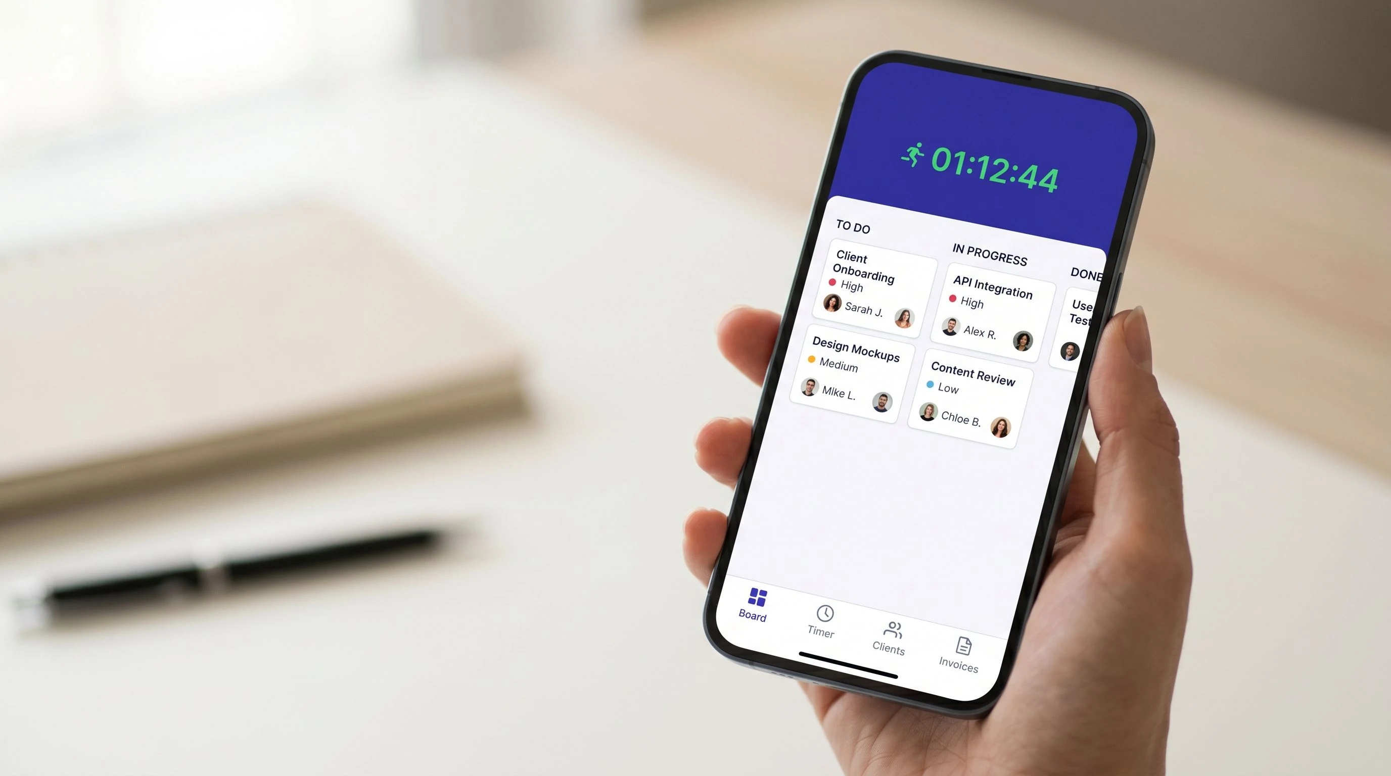 Smartphone with the app's project management dashboard — visible is a compact Kanban board with task cards and a running timer at the top of the screen, held by a hand over a light workspace surface